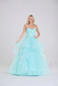 A woman wearing a voluminous mint green tulle ball gown stands in front of a pastel balloon wall. The dress features a sweetheart neckline, thin straps, a fitted embellished bodice, and a layered ruffle skirt.