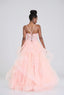 A woman stands with her back to the camera wearing a formal peach tulle ball gown with a lace-up back, posed in front of a pastel balloon wall.