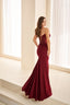 A woman modeling a strapless burgundy mermaid gown with lace-up back detailing, standing on a light-colored staircase in a bright, modern room with white curtains.